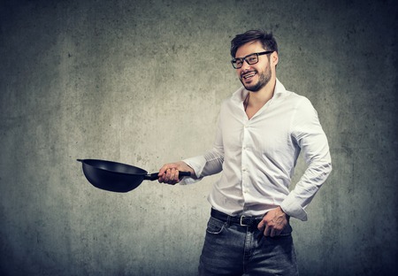 Young casual man holding frying pan and looking excited happy with cooking food on gray backgroundの写真素材