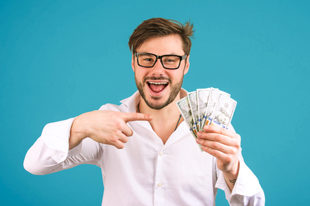 Cheerful successful man in white shirt holding heap of cash and pointing at it smiling at camera on blue backgroundの写真素材