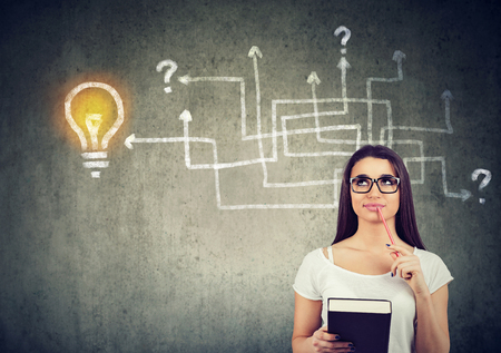 Woman with a book and pen brainstorming on new study project standing near a wall with arrow maze and a bright idea light bulb sketch on itの写真素材