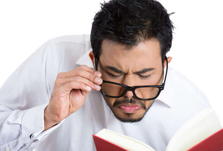 Closeup portrait of a young nerdy guy with big black eye glasses trying to read book but having difficulties seeing text because of vision problems.の写真素材