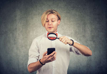 Portrait of a curious young man using mobile phone with magnifying glassの写真素材