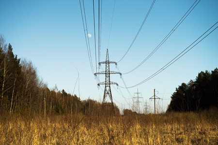 a line of high-voltage pillars through a plowed agricultural field, a forest on the horizon and a blue skyの写真素材