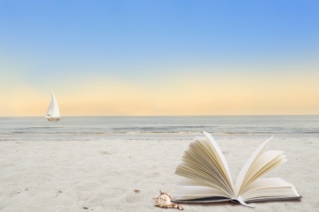 Lonely beach with book and shell in the foreground and sailboat in the background の写真素材