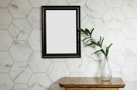 Elegant Mediterranean home interior. Summer home still life photo. Vase with green ruscus branches, wooden table. Blank picture frame mockup hanging on hexagon wallの写真素材