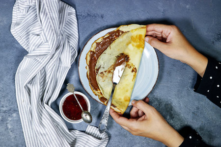 hands holding a plate of Crepes stuffed with chocolate spread on blue plate with fig jam bowl. Thin pancakes, blini. Sweet dessertの写真素材