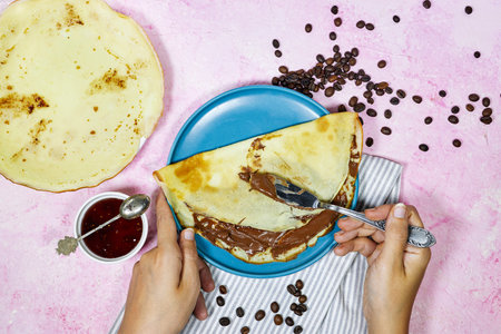 hands woman eating a Crepes stuffed with chocolate spread on blue plate with fig jam bowl and coffee beans on grunge pink background. Thin pancakesの写真素材