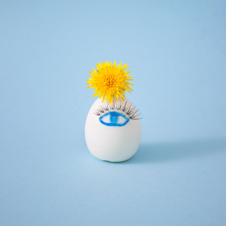 Creative white Easter egg with cute painted eye, eyelashes and yellow dandelion flower on pastel blue background. Happy Easter concept. Side view, interesting concept.の写真素材