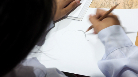 Background concept student girl has pencil in hand to sketching cartoon on the paper is her activity, Photo has her head foreground is blur and hand drawing is moving blur. On the wood desk has white book.の写真素材