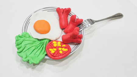 Molding clay breakfast sausage has egg, lettuce and tomato with a fork in ceramic plate, isolate on white.の写真素材