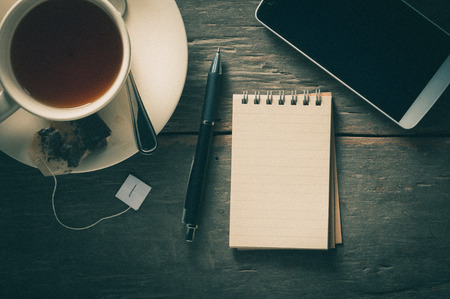 Small notepad with a cup of tea, pen, pencil and cellphne on rustic wood background with film filter...