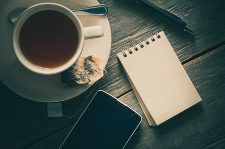 Small notepad with a cup of tea, pen, pencil and cellphne on rustic wood background with film filter effectの写真素材