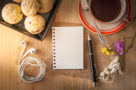 Opened notepad with pen, cookies in black ceramic dish, small earphone, and a cup of hot tea on wood table in cafe with morning light effectの写真素材