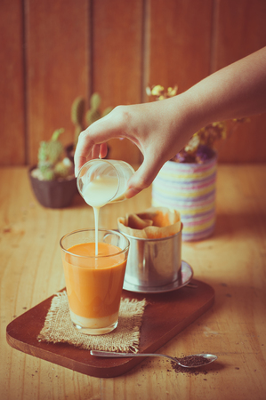 Woman right hand pouring fresh milk into glass of Dripped Thai tea by Vietnamese style on wood table in cafe with vintage filter effectの写真素材