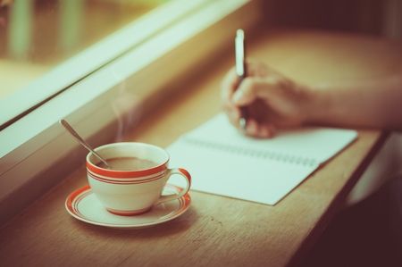 A coffee cup on wood bar beside window in cafe with male right hand writing on notebook in morning time with vintage film filter effectの写真素材