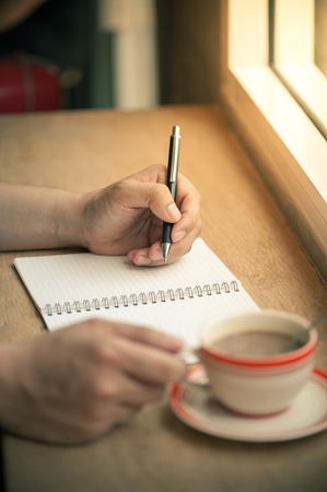 Male left hand writing on notebook with right hand holding coffee cup on wood bar beside window in cafe in morning timeの写真素材