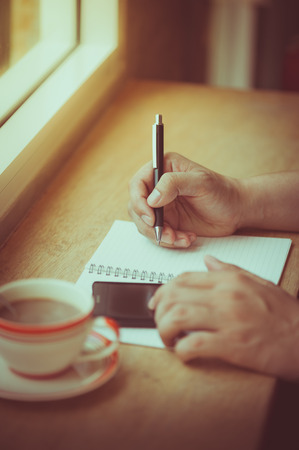 Male right hand writing on notebook with another hand using smartphone on wood bar beside window in cafe in morning time with vintage filter effectの写真素材