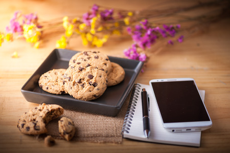 Chocolate chip cookies in black ceramic dish with smartphone, notebook and pen, and coffee cup at...