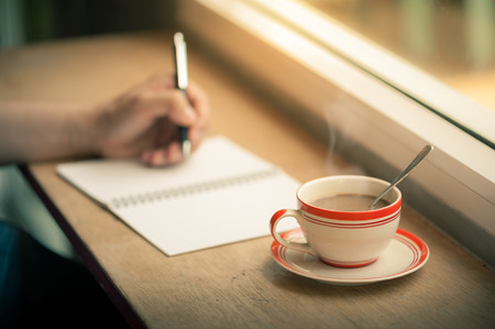 A coffee cup on wood bar beside window in cafe with male left hand writing on notebook in morning timeの写真素材