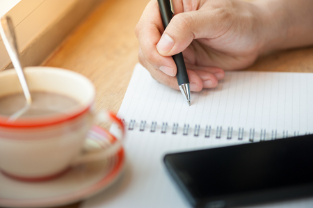 Male right hand writing on notebook with smart phone and coffee cup on wood bar beside window in cafe in morning timeの写真素材