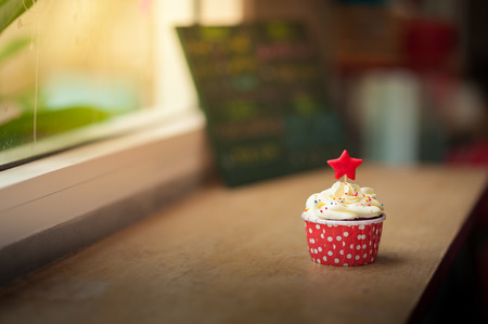 Small red velvet cupcake with cream that made from yogurt and cream cheese garnished with star shape fondant and colourful sugar sprinkle on wood bar in cafe in morning time with low key sceneの写真素材