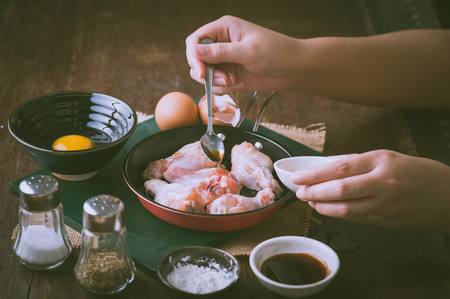 Preparation for cooking in home kitchen. Woman right hand pouring oyster sauce into fry pan to mix with chicken wings with vintage filter effect.の写真素材