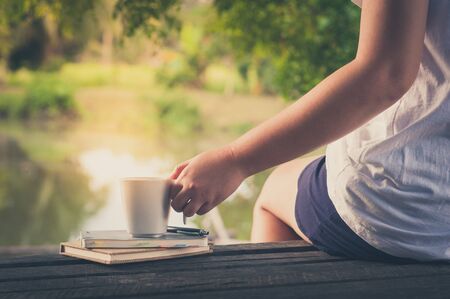 Woman left hand holding coffee cup beside notebooks on rustic wood bench with rural lake view in background in morning time on weekend with vintage filter effectの写真素材