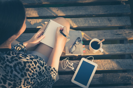 Young hipster woman writing journal on small notebook while sitting on wood bridge in morning time...