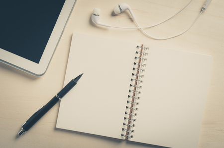 Notebook with blank area for text or message and pen beside small tablet pc and in ear headset on wood table with vintage filter effectの写真素材