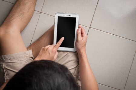 Young man with casual wear touching on small tablet pc screen while sitting on tile floor in his house on weekendの写真素材