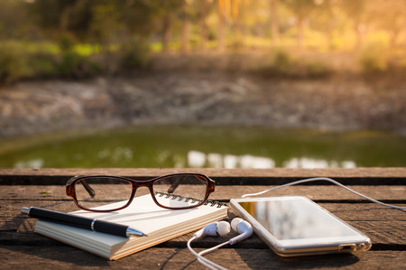 Smartphone with in ear headset, opened notebook, pen, and glasses on rustic wood table in morning time with natural lightingの写真素材