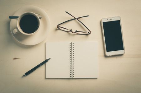 Opened notebook with blank area, pen, smart phone, and coffee cup on wood table in morning time with vintage filter effectの写真素材