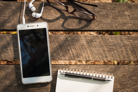 Smartphone with blank area on touchscreen with in ear headset, opened notebook, pen, and glasses on rustic wood table in morning time with natural lightingの写真素材