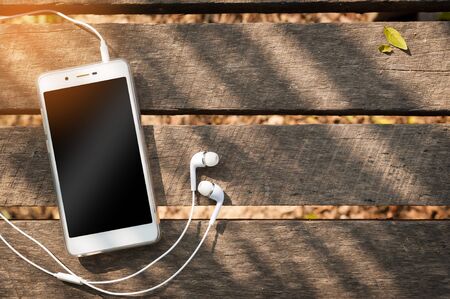 Smartphone with blank area on touch screen and in ear headphone on rustic wood table at outdoor area in morning time with high contrast natural lightingの写真素材