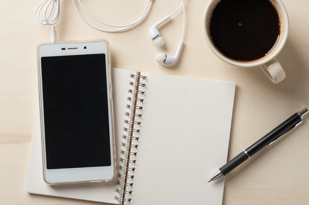 Smartphone with blank area on touchscreen with in ear headset, notebook, pen, glasses, and coffee cup on wood tableの写真素材