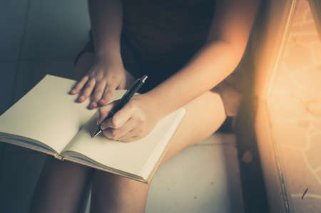 Young woman left hand writing on blank book while sitting beside window in morning time on weekend....