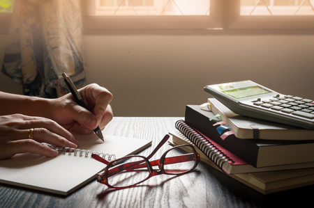 Male left hand writing on notebook beside glasses and books on table in morning time on work day. Freelance business working concpetの写真素材