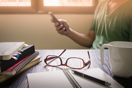 Glasses put down on table beside notebooks and pen while young freelance worker using smart phone on blurry background in morning time on work day. Working at home conceptの写真素材