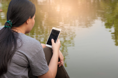 Weekend morning lifestyle. Young woman touching on mobile phone screen while sitting outdoor beside river in morning time. Freelance working and phone addiction conceptの写真素材