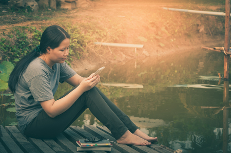 Weekend morning lifestyle. Young woman using her mobile phone seriously while sitting outdoor beside lake in morning time. Freelance working and phone addiction concept with vintage filter effectの写真素材