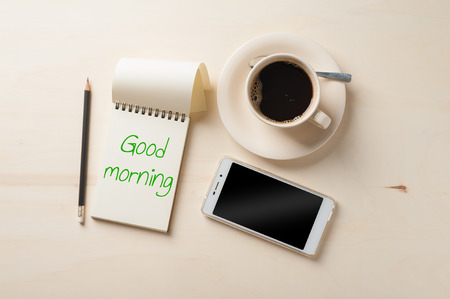 "Good morning" is written on opened notebook with blank area, pen, smart phone, and coffee cup on wood table in morning timeの写真素材