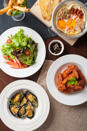 Top view of modern cuisine set including spring rolls, pan fried egg, Korean style deep fried chicken, baked mussels, and Japanese crab stick salad on wood table in restaurantの写真素材