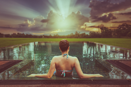 Vacation lifestyle scene of young woman sitting in swimming pool in morning time with sunset landscape view in blurry background. Weekend and holiday lifestyle concept with vintage filter effectの写真素材