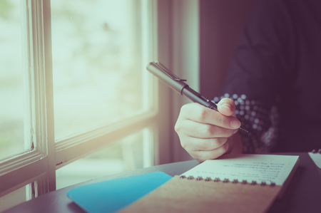 Morning scene of young hipster woman right hand writing on small notebook beside window in cafe. Freelance working lifestyle with vintage filter effectの写真素材