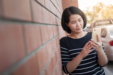 Weekend activity scene of adult Asian woman using mobile phone while standing beside brick wall. Urban lifestyle on with technology.の写真素材