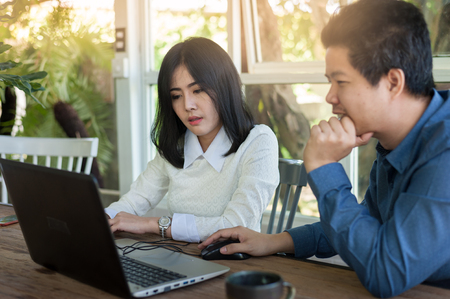Young Asian businessman and businesswoman talking about their work while looking at laptop monitor in coffee shop. Startup business for freelance workers conceptの写真素材