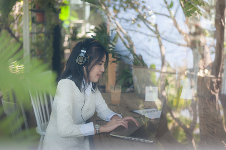 Young attractive Asian woman listening music while working online with laptop computer in coffee shop. Freelance worker and self-employed business activity conceptの写真素材
