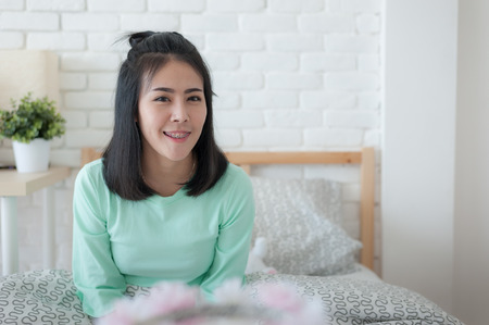 Young attractive Asian woman wear green casual cloth smiling while sitting on bed. Lazy girl activity on sunday morning.の写真素材