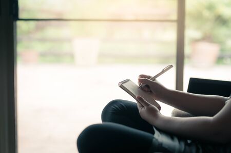 Woman hand writing some idea on small notepad while sitting on chair in house. Freelance working from anywhere concept with vintage filter effectの写真素材