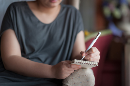 Left-handed woman writing some idea on small notepad while sitting on chair in house. Freelance working from anywhere conceptの写真素材