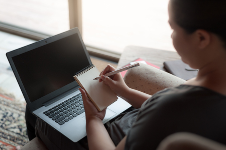 Woman freelancer writing memo on small notepad while working with laptop computer. Online business and self-employed conceptの写真素材